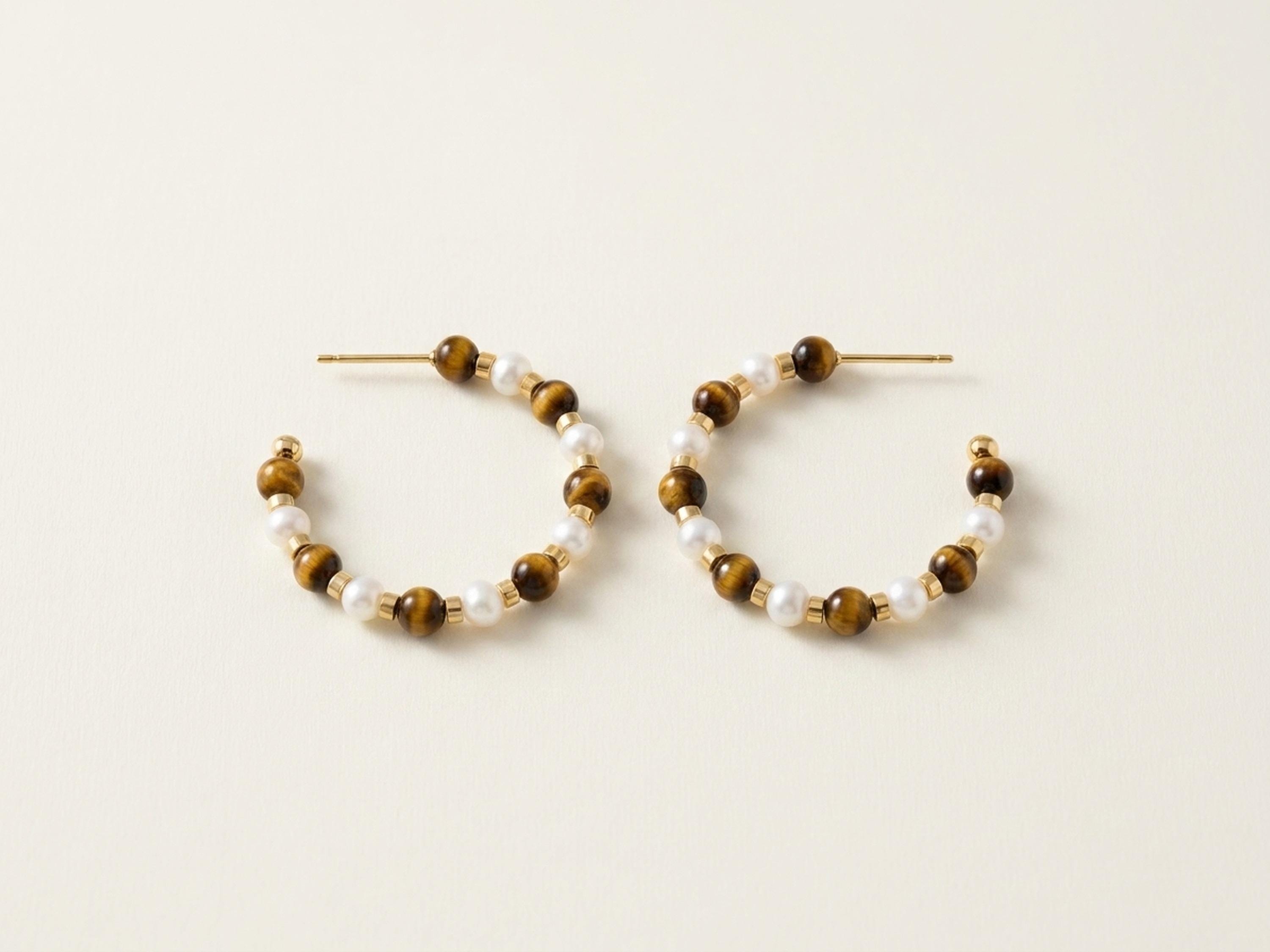 Pair of tiger eye and white pearl beaded hoop earrings on a neutral cream background