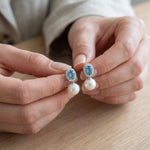 Hands holding matching blue oval cubic zirconia and white pearl drop earrings