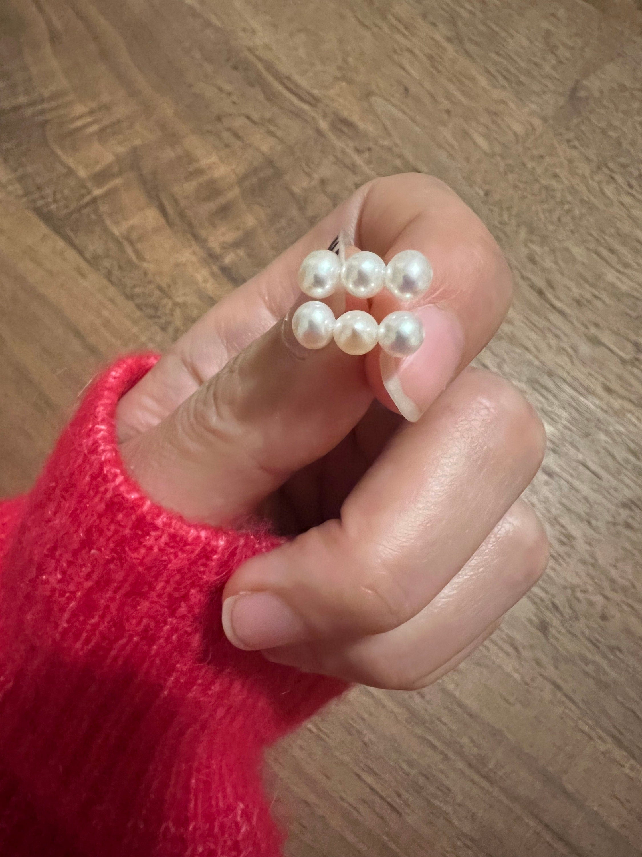 A hand in a red sweater holds two three-pearl earrings.