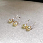 Four golden freshwater pearl stud earrings are displayed on a light gray surface.