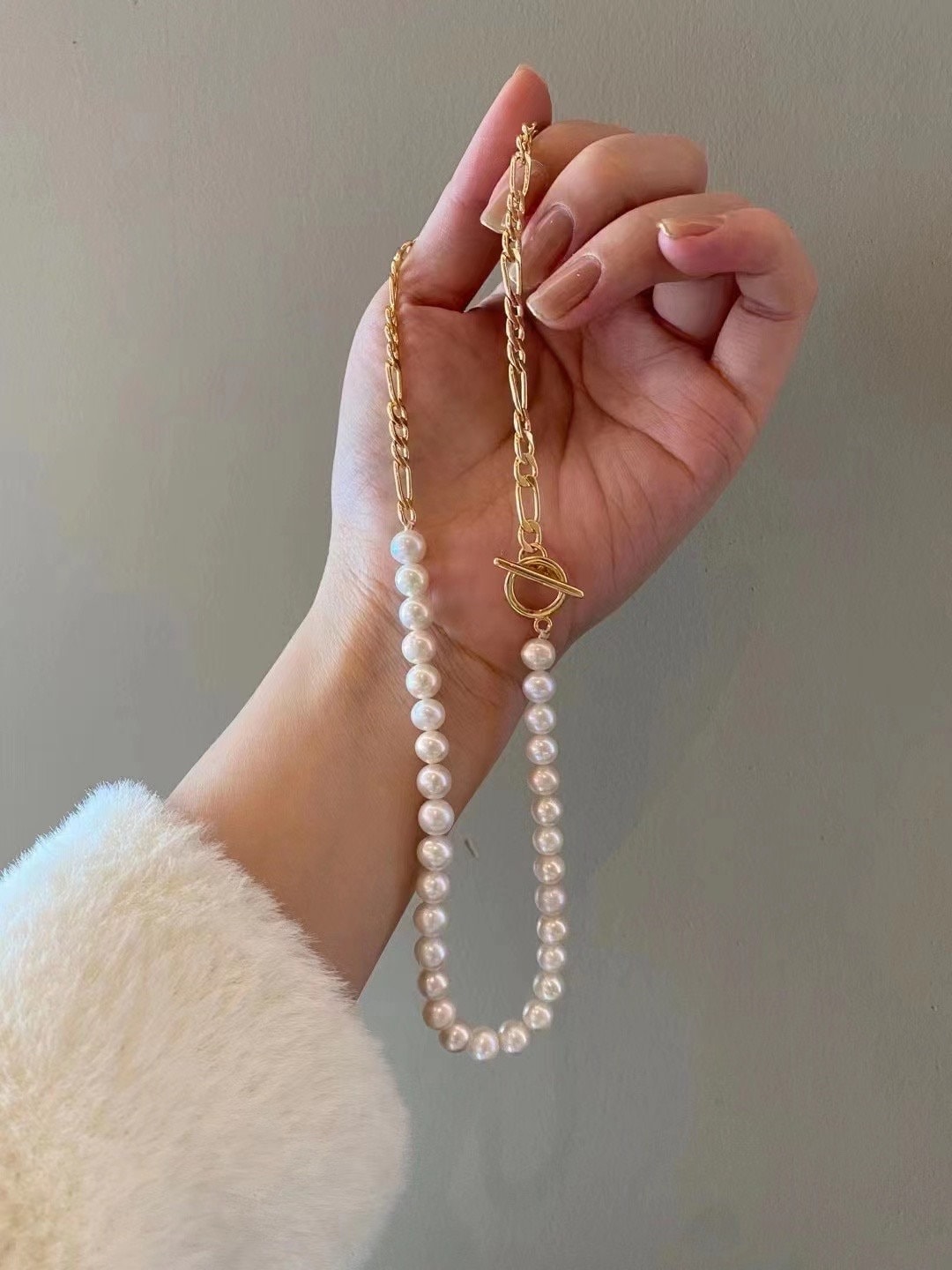 A hand holds a freshwater pearl necklace with a gold-plated chain and toggle clasp.