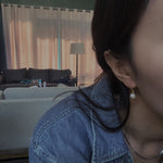 A woman wears a single pearl hoop earring, with a denim shirt and dark hair.