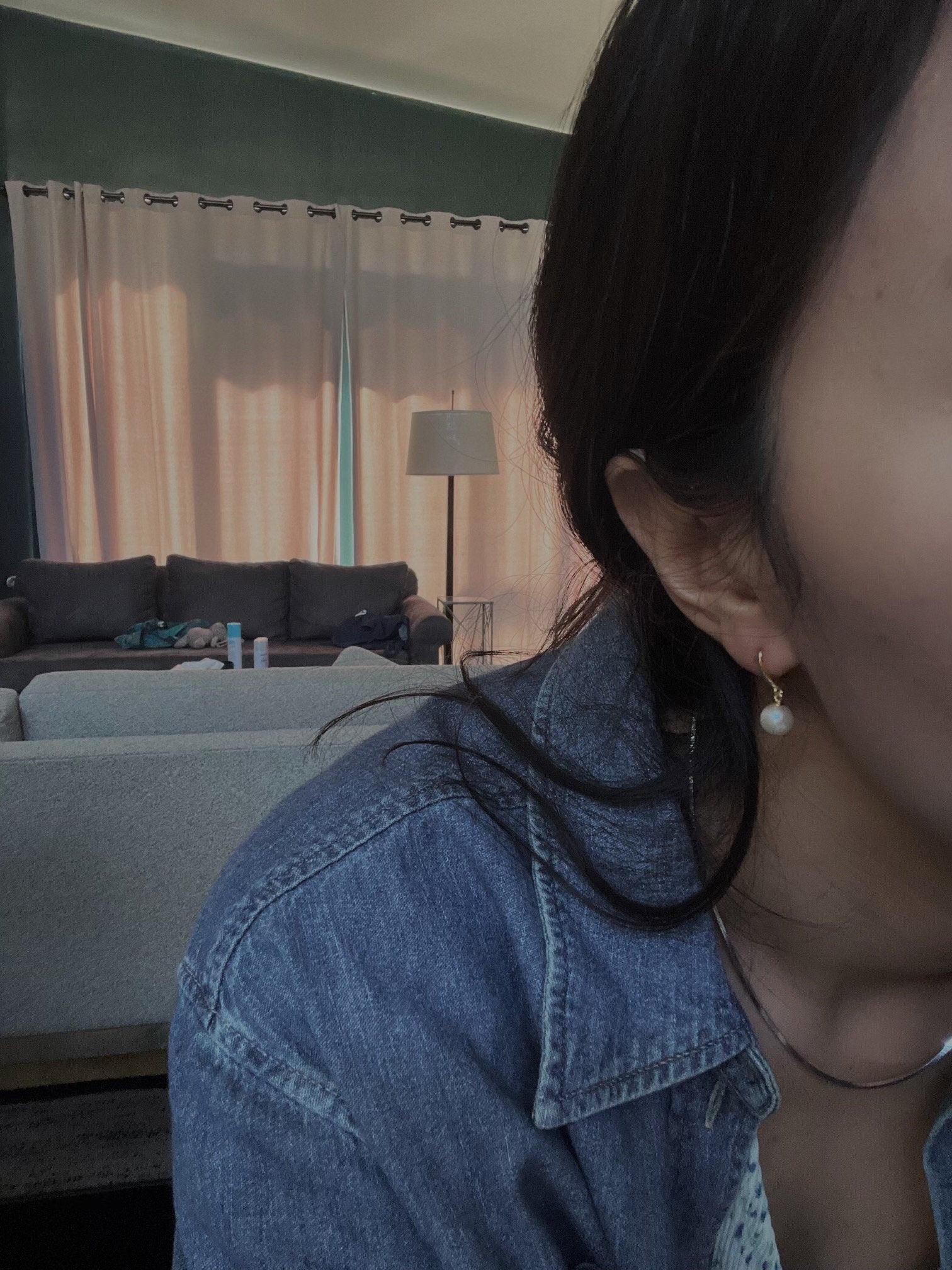 A woman wears a single pearl hoop earring, with a denim shirt and dark hair.