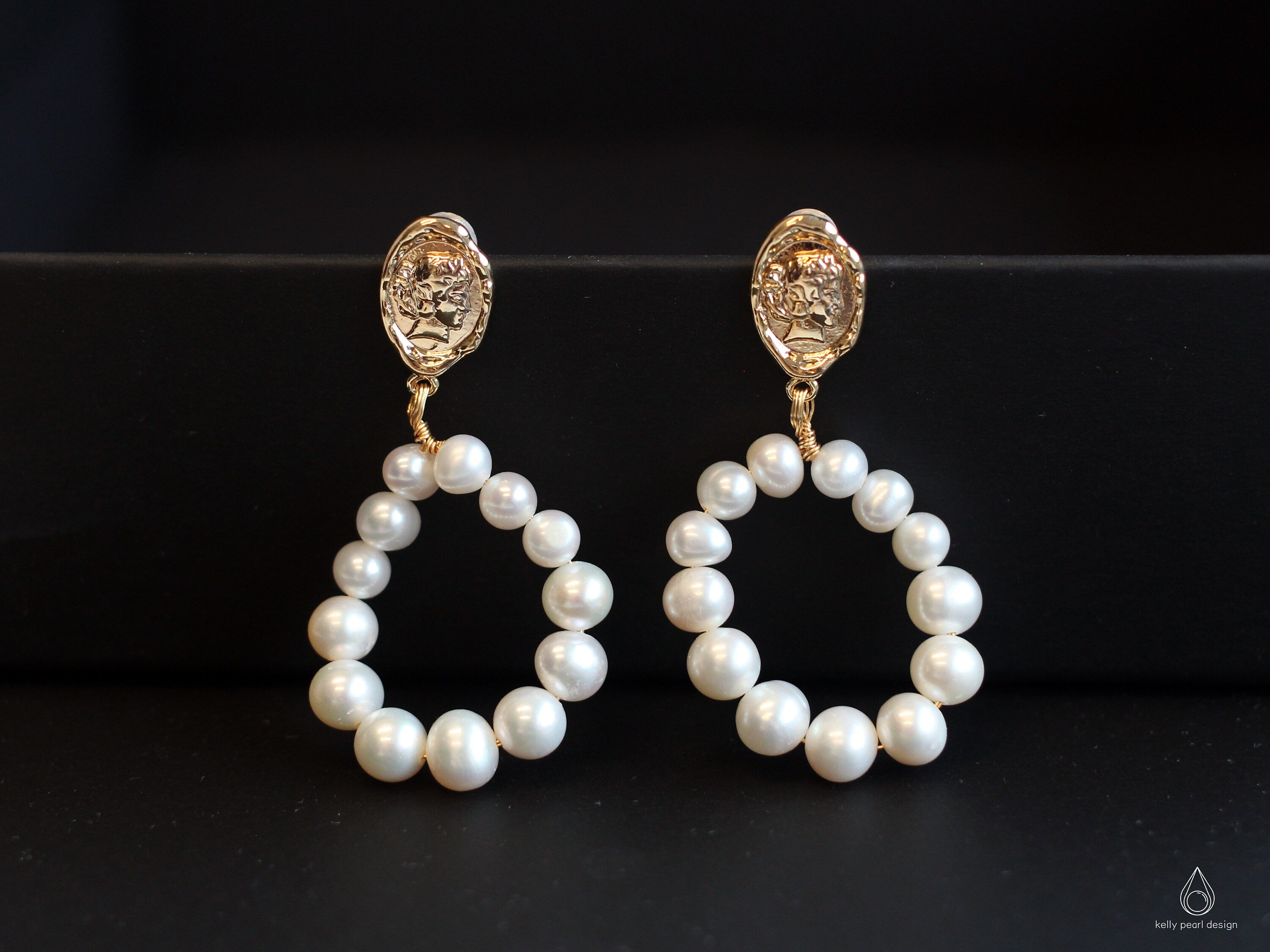 Two gold and freshwater pearl hoop earrings are displayed on a black surface.