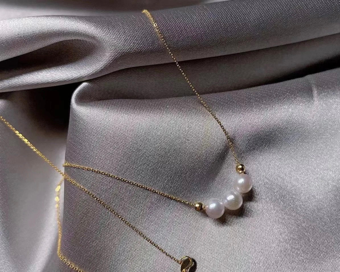 A delicate gold chain necklace featuring three white pearls, displayed on a silver-grey fabric.