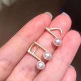 Two gold-plated musical note stud earrings, each with a freshwater pearl, held in a hand.