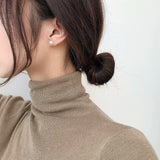 A woman wears a single pearl stud earring and a tan turtleneck sweater.